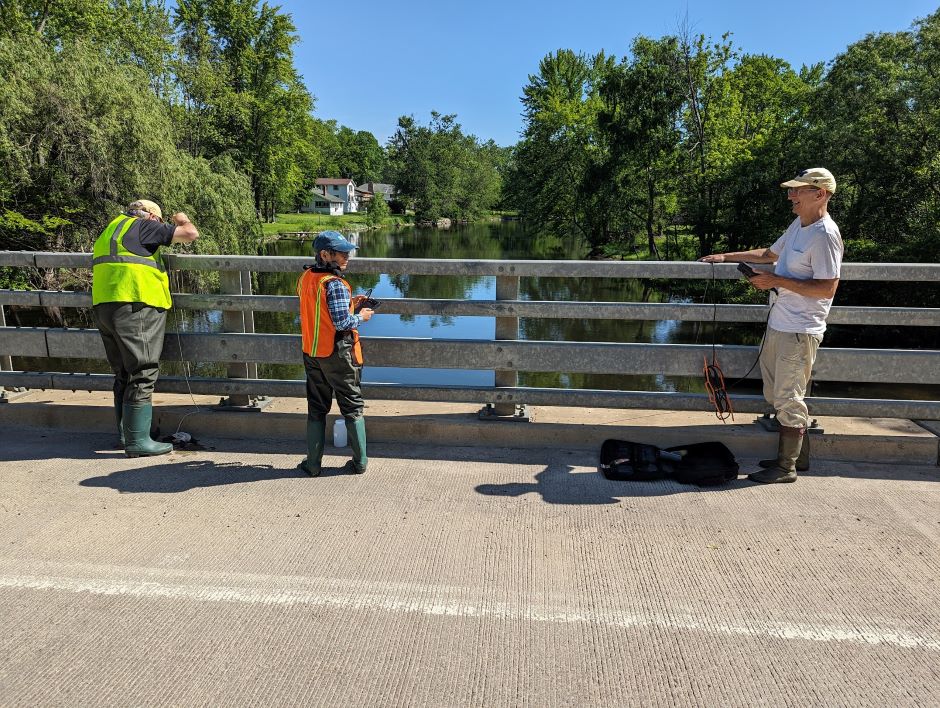 Three experienced HRWC Chemistry and Flow Monitoring citizen science volunteers work together to record YSI parameter data and collect additional water samples by lowering a Van Dorn Sampler into the main stem of the Huron River off a road overpass south of Brighton, Michigan. 